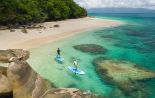 Two people paddleboard in clear turquoise water beside a tropical beach lined with rainforest and granite boulders.